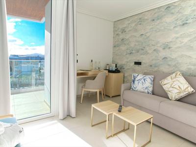 Helles Wohnzimmer mit grauer Couch, zwei kleinen Tischen, Schreibtisch und Balkon mit Ausblick.