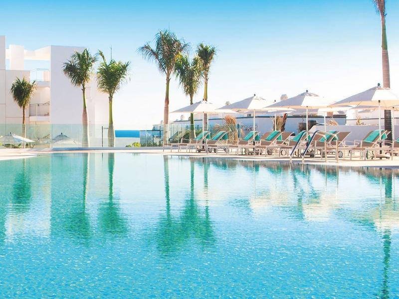 Großer Pool mit klarem blauen Wasser, Sonnenliegen und Palmen vor weißem Gebäude und blauem Himmel.