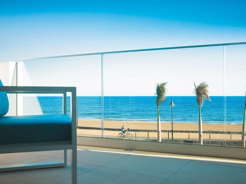 Blick auf das Meer von einer Terrasse mit Glasgeländer, blauer Stuhl links und Palmen am Strand.