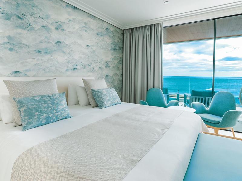 Helles Schlafzimmer mit großem Bett, blau-weißer Tapete und Meerblick durch bodentiefe Fenster.