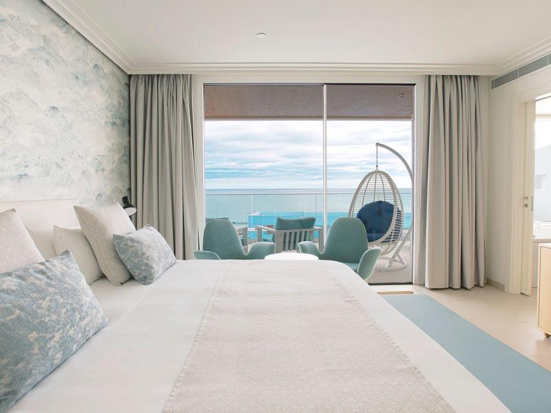 Helles Hotelzimmer mit großem Bett, Fenster mit Meerblick und Hängesessel auf Balkon.