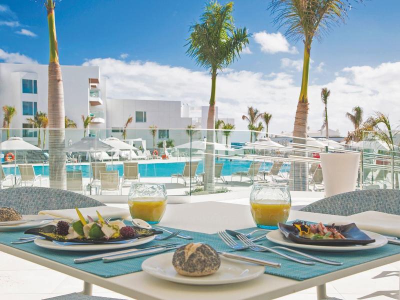Modernes Außenrestaurant mit Pool, Palmen, blauem Himmel und gedecktem Tisch mit Getränken.