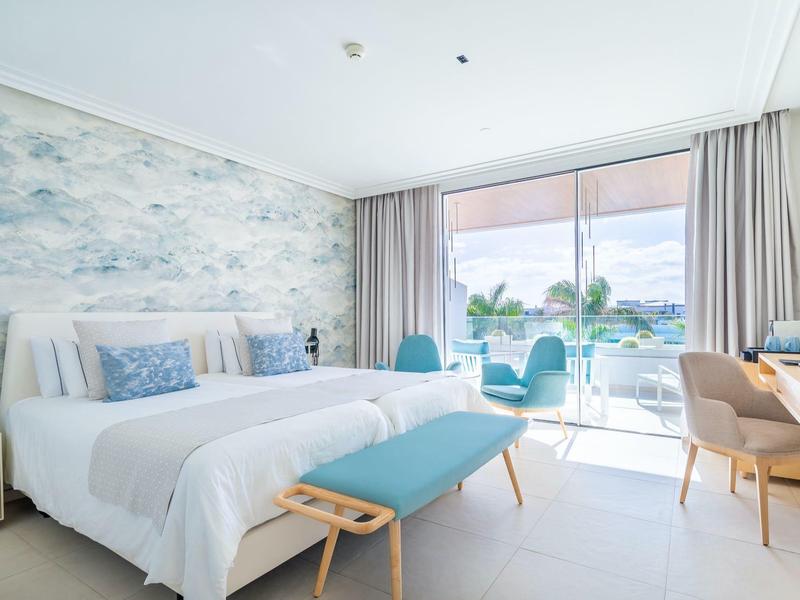 Helles Hotelzimmer mit großem Bett, blauer Bank, Schreibtisch und Balkon mit Meerblick bei Tageslicht.