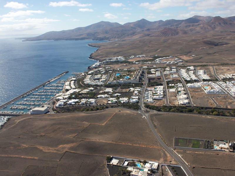 Küstenstadt mit Hafen, weißen Gebäuden, Bergen im Hintergrund und trockenem, braunem Gelände.