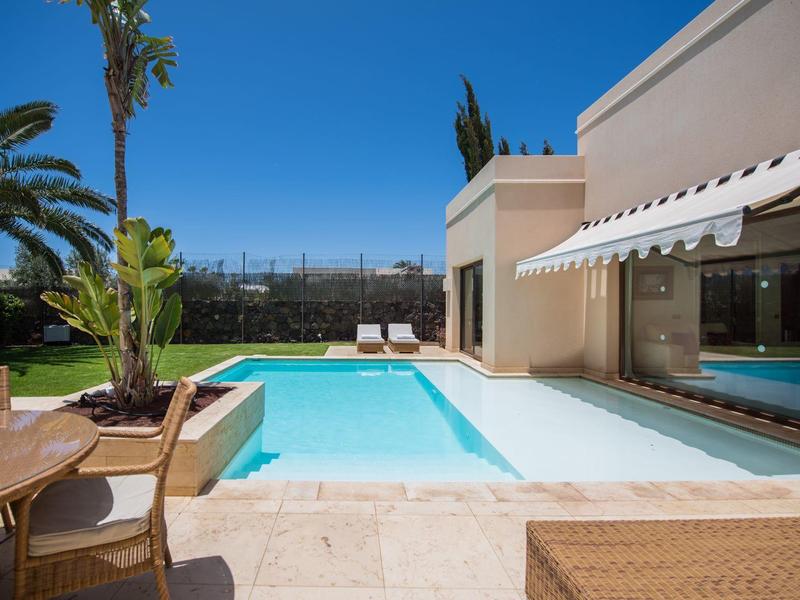 Moderner Poolbereich mit klarem Wasser, Liegestühlen, Palmen und Sandsteinboden an sonnigem Tag.