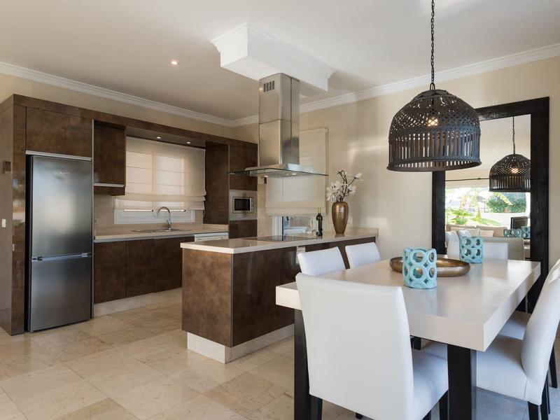 Moderne Küche mit dunklen Holzschränken, weißem Esstisch, sechs weißen Stühlen und einem Hängeleuchter.