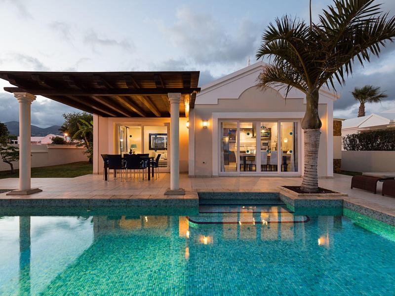 Haus mit Pool, Terrasse, Palmbaum und beleuchtetem Außenbereich bei Dämmerung.