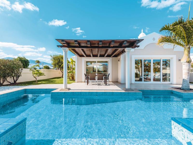 Moderner weißer Bungalow mit großem Pool, Terrasse, Pergola und Palmen in sonniger Landschaft.