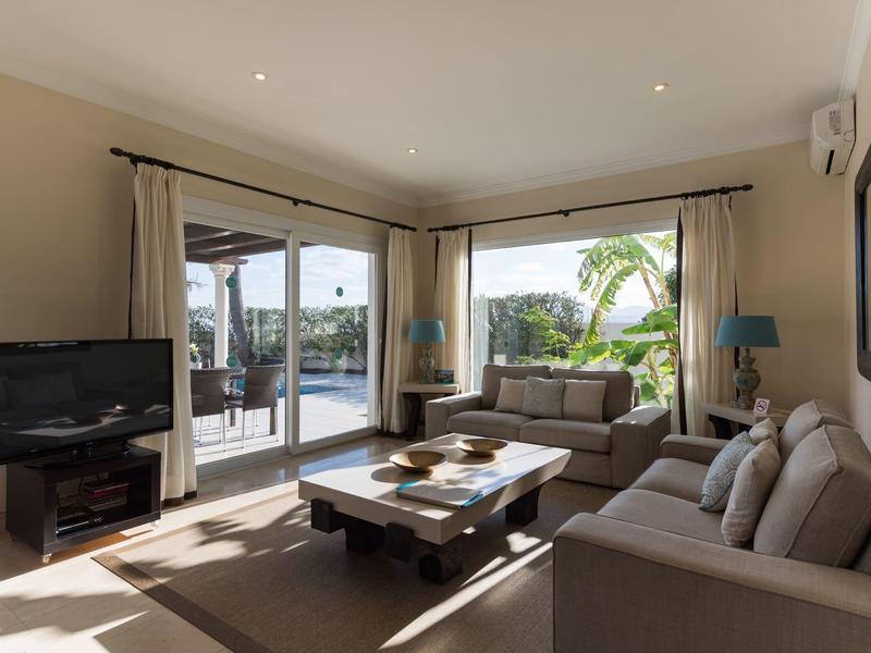 Heller Wohnraum mit grauen Sofas, großem Holztisch, TV und Blick auf Terrasse mit Pool.