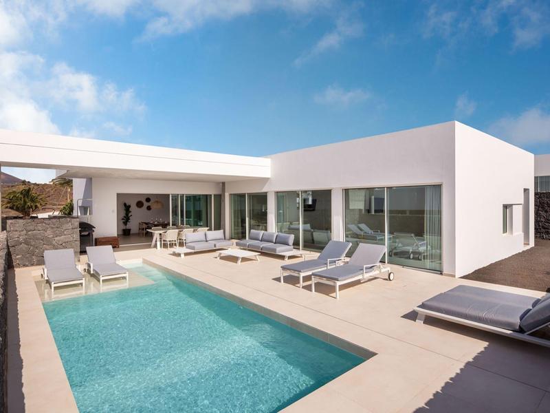 Moderner weißer Bungalow mit Pool, Liegestühlen und Glasfront unter blauem Himmel.