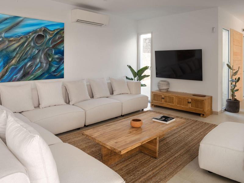 Helles Wohnzimmer mit weißen Sofas, Holztisch, großem Wandbild und TV auf hellbraunem Teppich.