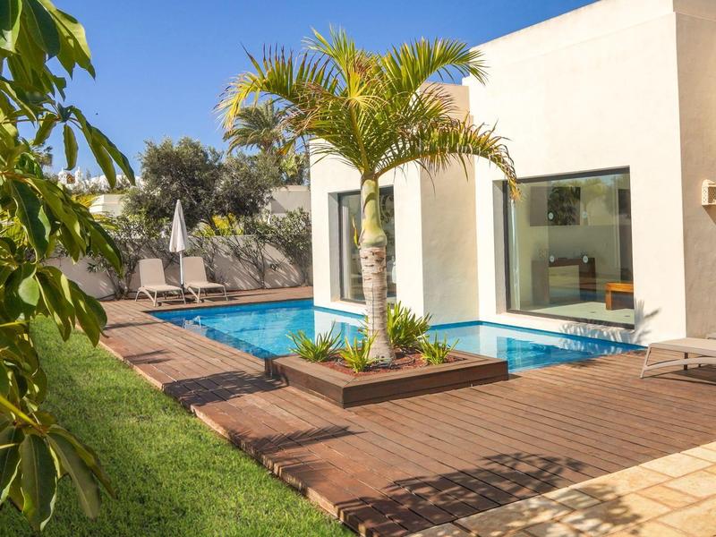 Moderner Außenbereich mit kleinem Pool, Holzterrasse, Palmen und Liegestühlen bei klarem Himmel.