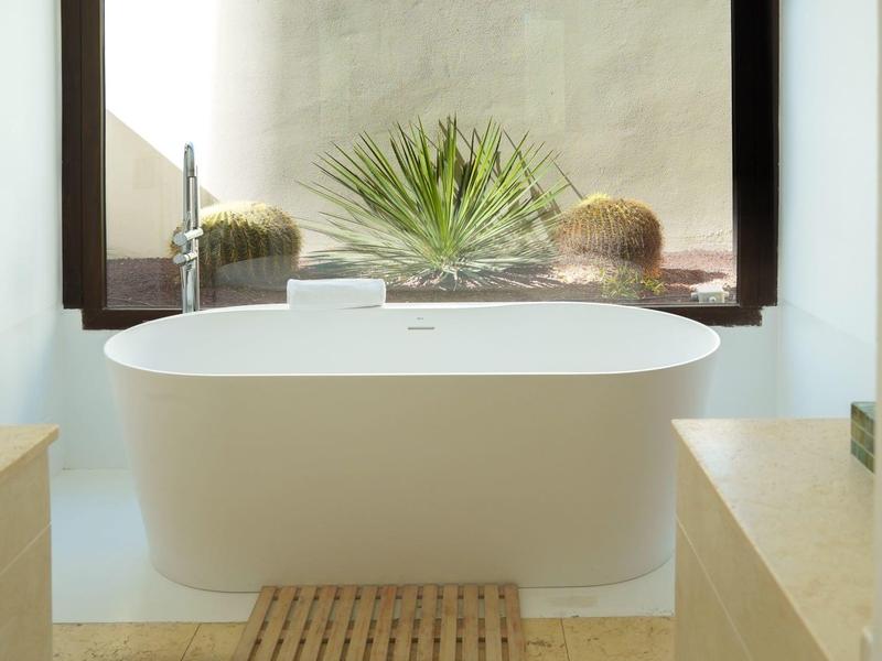 Moderne weiße Badewanne vor großem Fenster mit Pflanzen, beige Fliesen und Holzmatte.