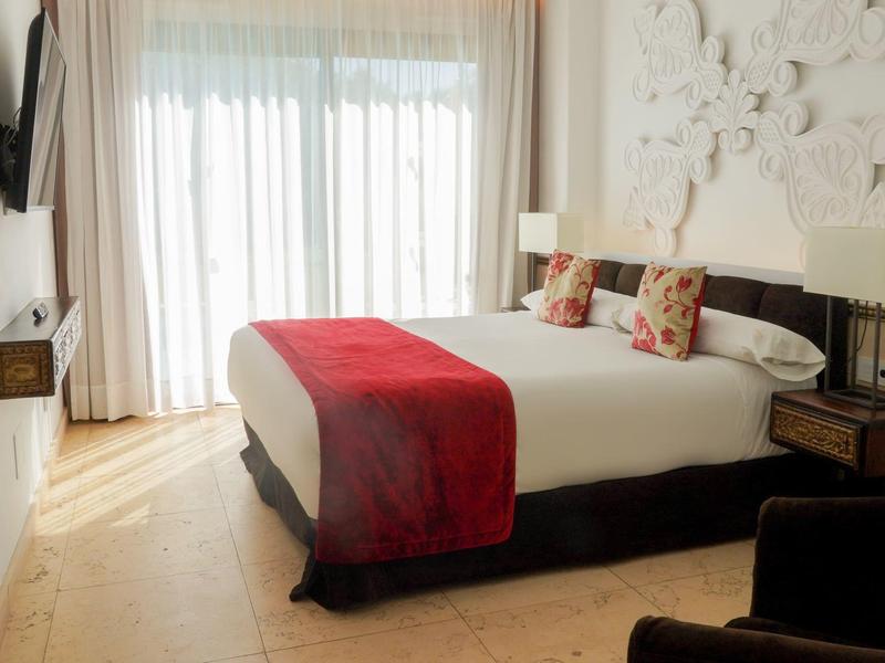 Modernes Hotelzimmer mit großem Bett, rotem Überwurf, weißen Vorhängen und hellem Holzboden.