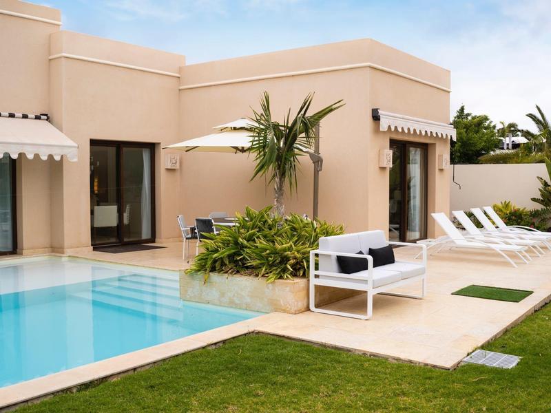 Moderner Garten mit rechteckigem Pool, beigem Haus, weißen Sonnenliegen und grünem Rasen.