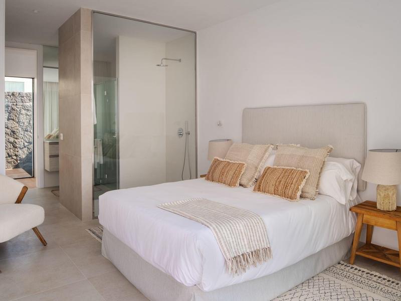 Modernes Schlafzimmer mit weißem Bett, Holznachttisch, Lampe und Stuhl, heller Raum mit Glasdusche.