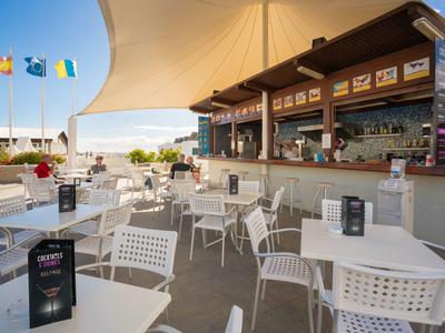 Außenbar mit weißen Tischen und Stühlen unter Sonnensegel, bunte Fahnen im Hintergrund.