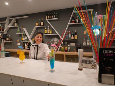 Frau hinter weißer Bar mit bunten Cocktails, Regale mit Flaschen und farbige Trinkhalme im Hintergrund.