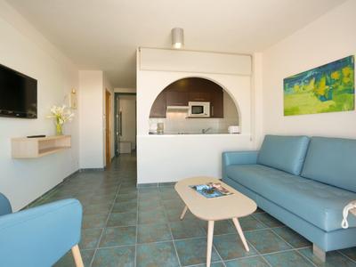 Helles Wohnzimmer mit blauer Couch, kleinem Tisch, Wand-TV und Küchenöffnung mit Mikrowelle.