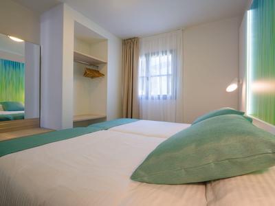 Ein einfaches Schlafzimmer mit großem Bett, grünem Kissen, offenen Schrank und Fenster mit Vorhängen.