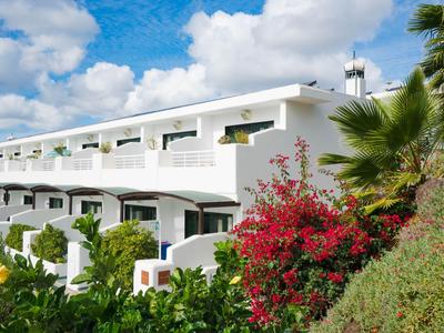 Bâtiment hôtelier blanc avec balcons entouré de plantes vertes et de fleurs rouges sous un ciel bleu.