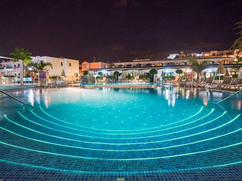 Großer beleuchteter Pool bei Nacht mit umliegenden weißen Gebäuden und Palmen.