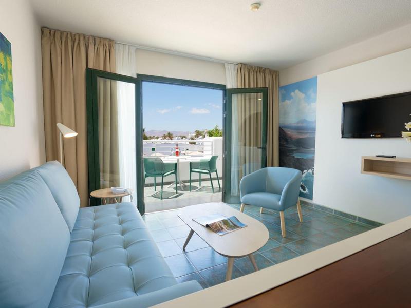 Hell eingerichtetes Wohnzimmer mit hellblauem Sofa, Sesseln und offener Balkontür mit Stadtblick.