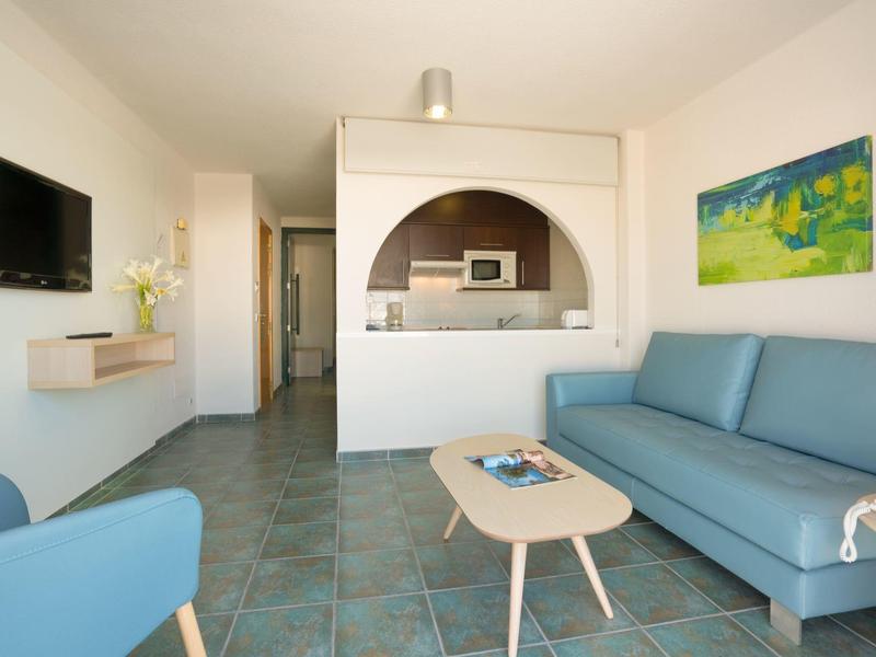 Helles Wohnzimmer mit blauer Couch, kleinem Tisch, Wand-TV und Küchenöffnung mit Mikrowelle.