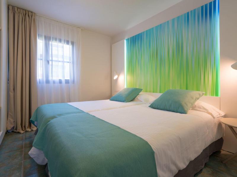 Hell eingerichtetes Schlafzimmer mit Doppelbett, grüner Tagesdecke, Kissen und farbigem Wandbild.