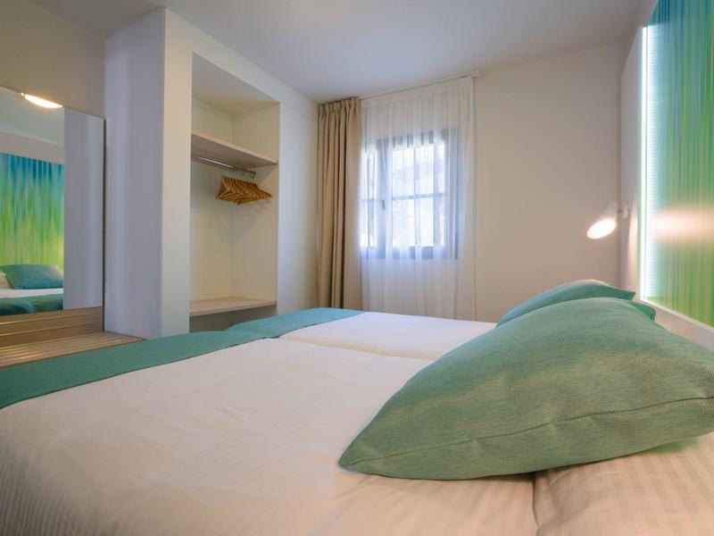 Ein einfaches Schlafzimmer mit großem Bett, grünem Kissen, offenen Schrank und Fenster mit Vorhängen.