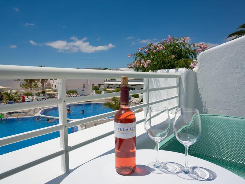 Flasche Roséwein und zwei leere Gläser auf weißem Tisch auf Balkon mit Pool und blauem Himmel.