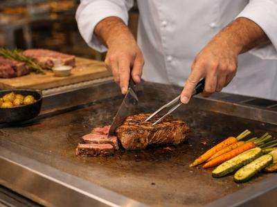 Koch wendet Steak und grillt Gemüse auf einer großen Grillfläche.