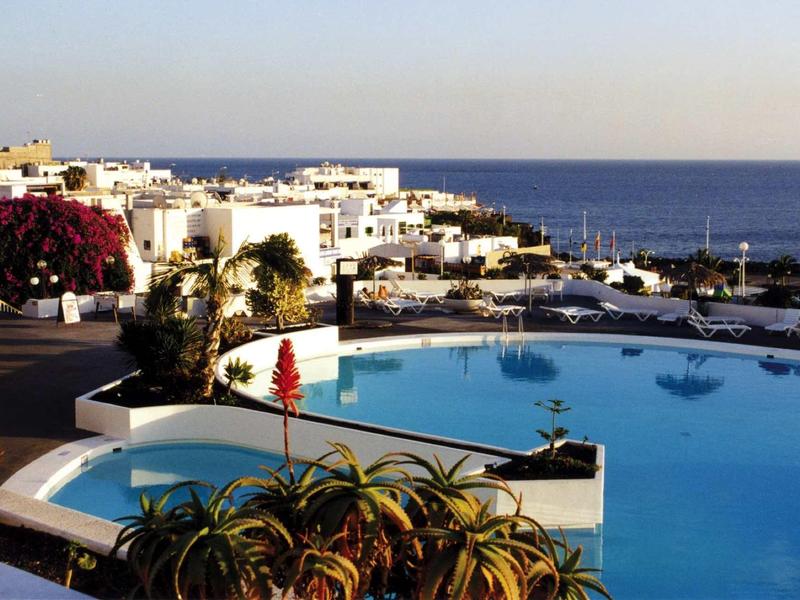 Blick auf einen Pool mit weißen Gebäuden und Meer im Hintergrund bei klarem Himmel.