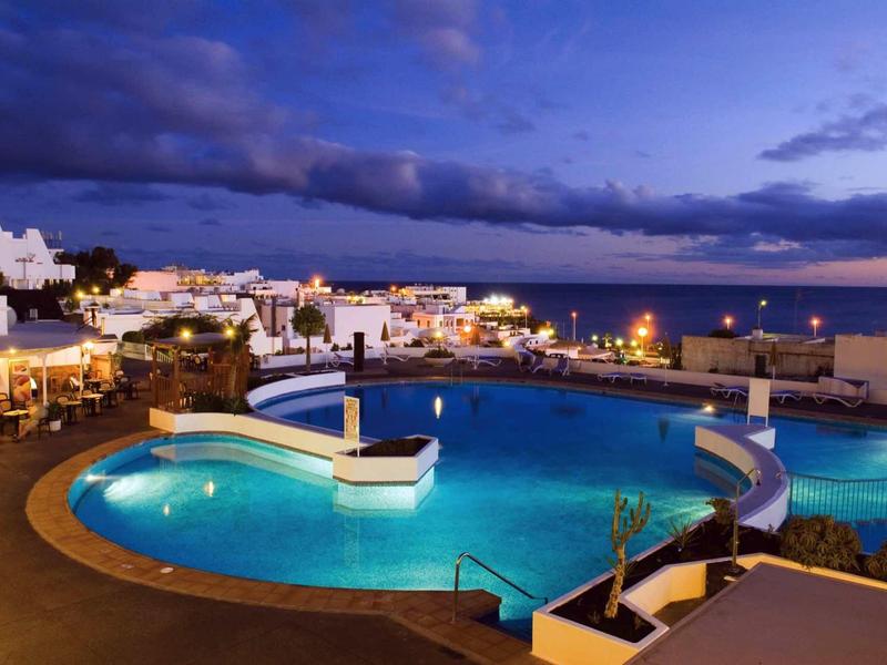 Beleuchteter runder Pool mit Sitzbereich, dahinter hell erleuchtete Häuser und ein dunkler Abendhimmel.