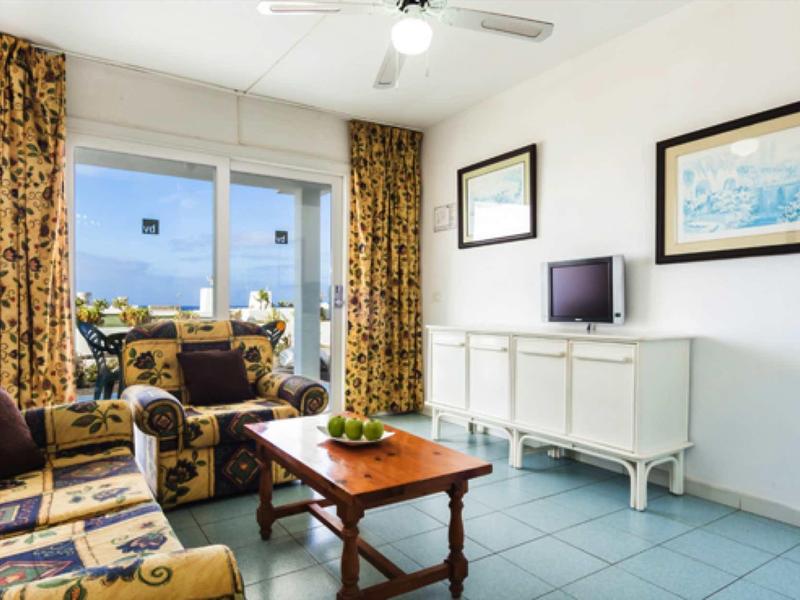 Helle, gemütliche Wohnzimmer mit blau-gelber Polstermöblierung, Holztisch und weißen Möbeln.