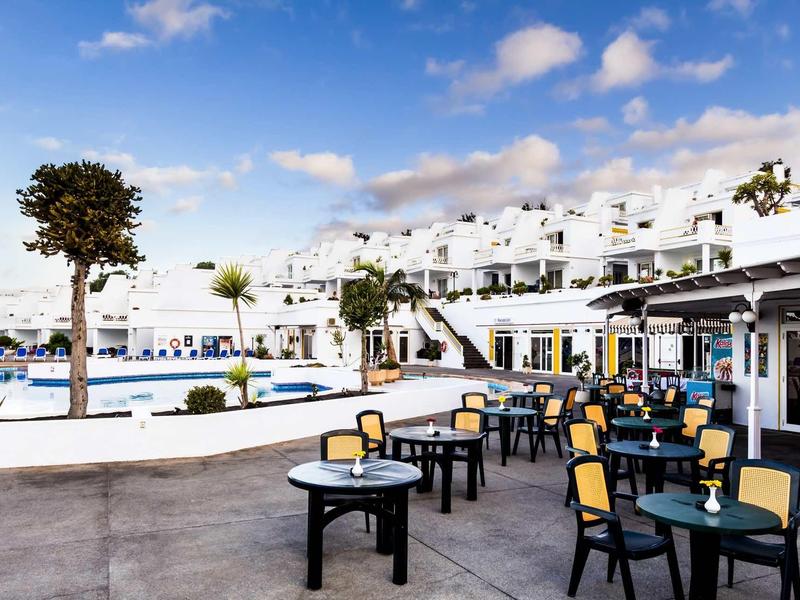 Strandbar mit schwarzen Tischen und Stühlen, weißem Gebäude und blauem Himmel mit Wolken.