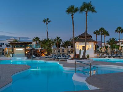 Area piscina illuminata dell'hotel con lettini e palme al crepuscolo.