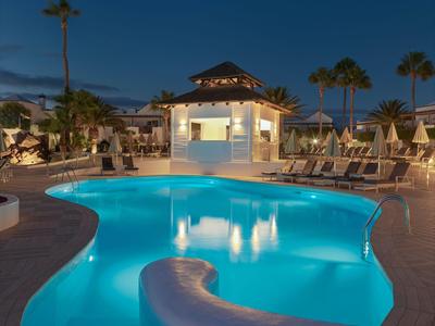 Beleuchteter Poolbereich mit Liegestühlen und Palmen bei Nacht in einem Resort.
