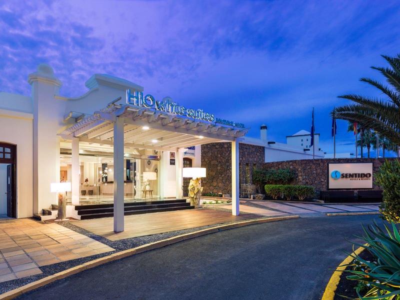 Eingang eines modernen Hotels mit heller Beleuchtung, Palmen und blauem Abendhimmel.