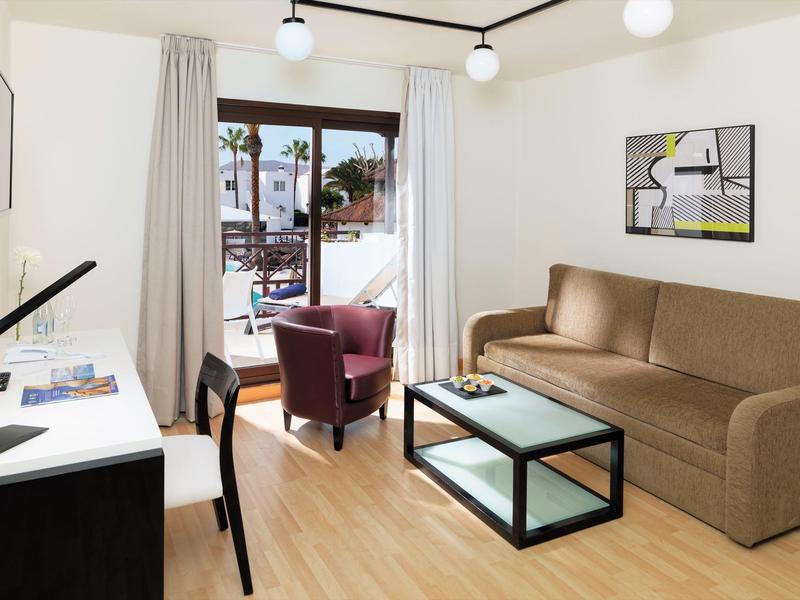 Modernes Wohnzimmer mit beigem Sofa, Glastisch, zwei Sessel und hellem Holzboden, Balkon mit Blick.