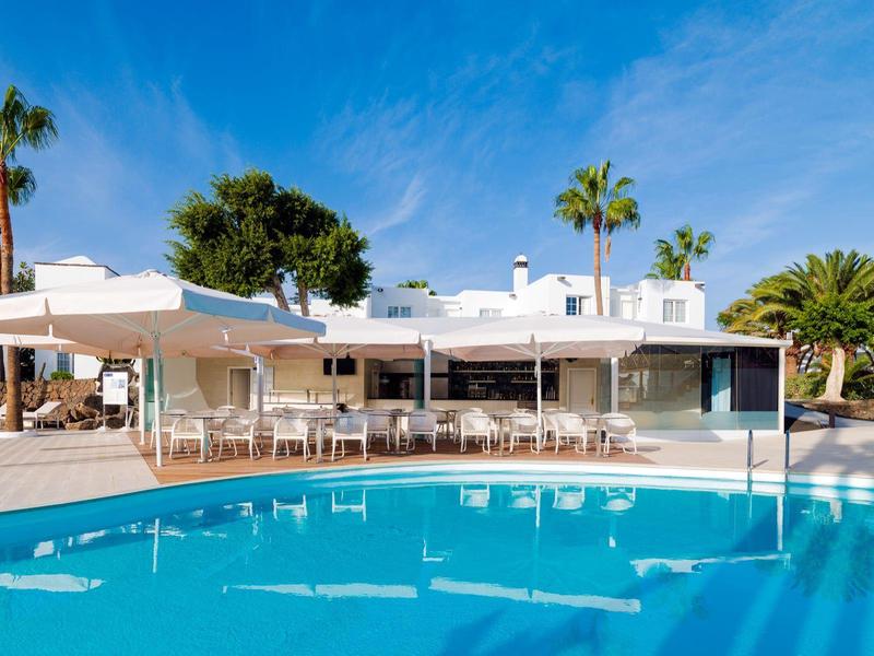 Moderner weißer Bungalow mit großem blauen Pool und Palmen bei klarem Himmel.