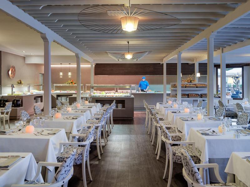 Großer heller Speisesaal mit weißen gedeckten Tischen, Holzstühlen und Deckenlampen.