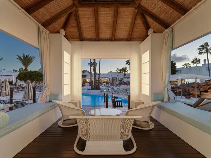 Moderner Poolbereich mit Lounge, weißem Mobiliar und Panoramafenstern mit Blick auf Palmen.