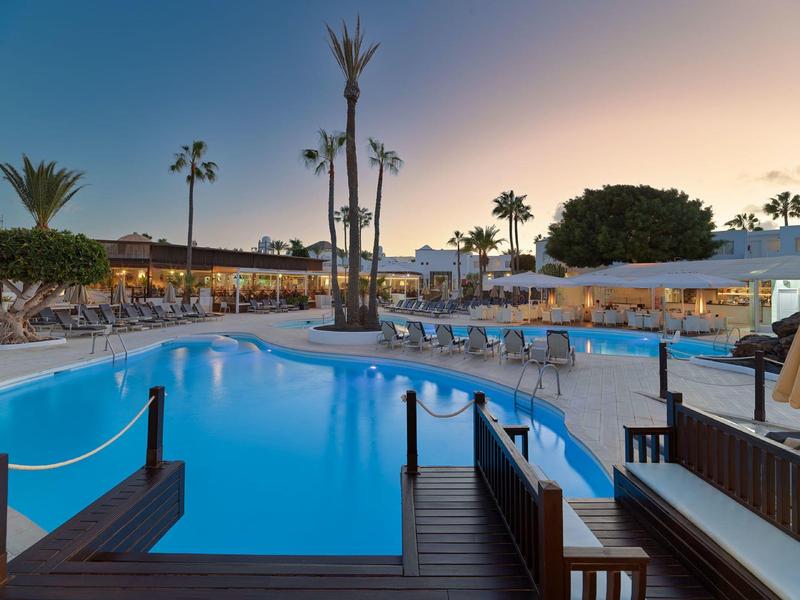 Poolbereich mit blauer Wasserfläche, Liegestühlen und Palmen bei Sonnenuntergang.