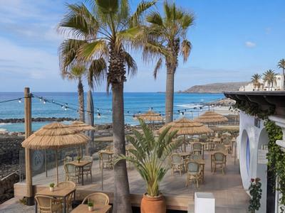 Strandterrasse mit Tischen, Stühlen und Palmen vor blauem Meer und Himmel.