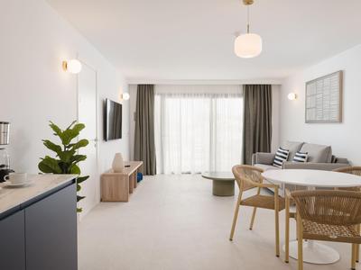 Helles Wohnzimmer mit Esstisch, Sofa und Fernseher neben bodentiefen Fenstern.