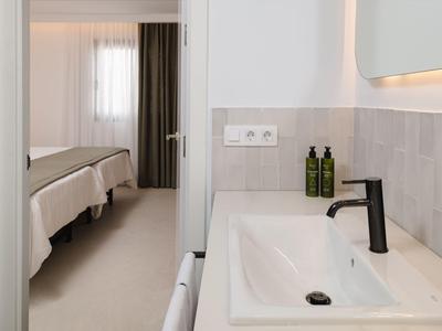 Hell eingerichtetes Hotelzimmer mit Badezimmer, weißem Waschbecken und Blick auf das Schlafzimmer.