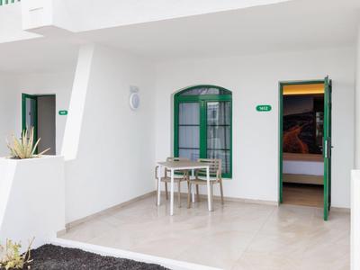 Weißes Haus mit grünen Türen, Terrasse mit kleinem Tisch und zwei Stühlen, zwei Topfpflanzen.