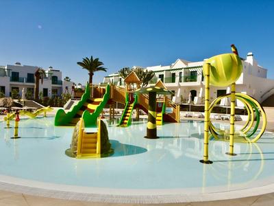 Kinder-Wasserspielplatz mit grünen Rutschen und gelben Wasserspielen vor weißen Wohnungen.