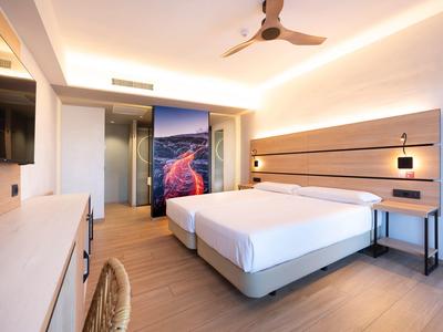 Modernes Hotelzimmer mit großem Bett, Ventilator an der Decke und Stuhl aus geflochtenem Material.